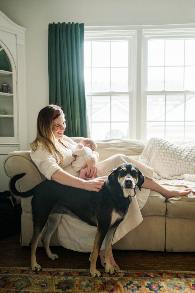 Mom holds newborn baby while petting dog in her living room for her Louisville in-home newborn session.