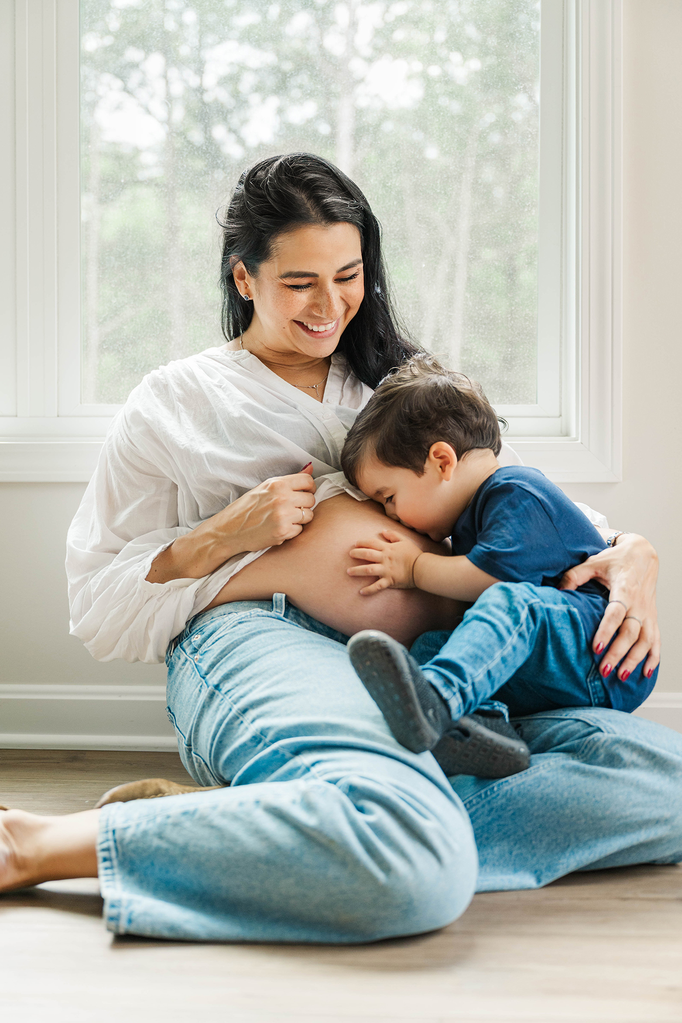 Pregnant mother sitting by a window as her toddler son kisses her belly, a favorite Louisville Maternity Photographer pose