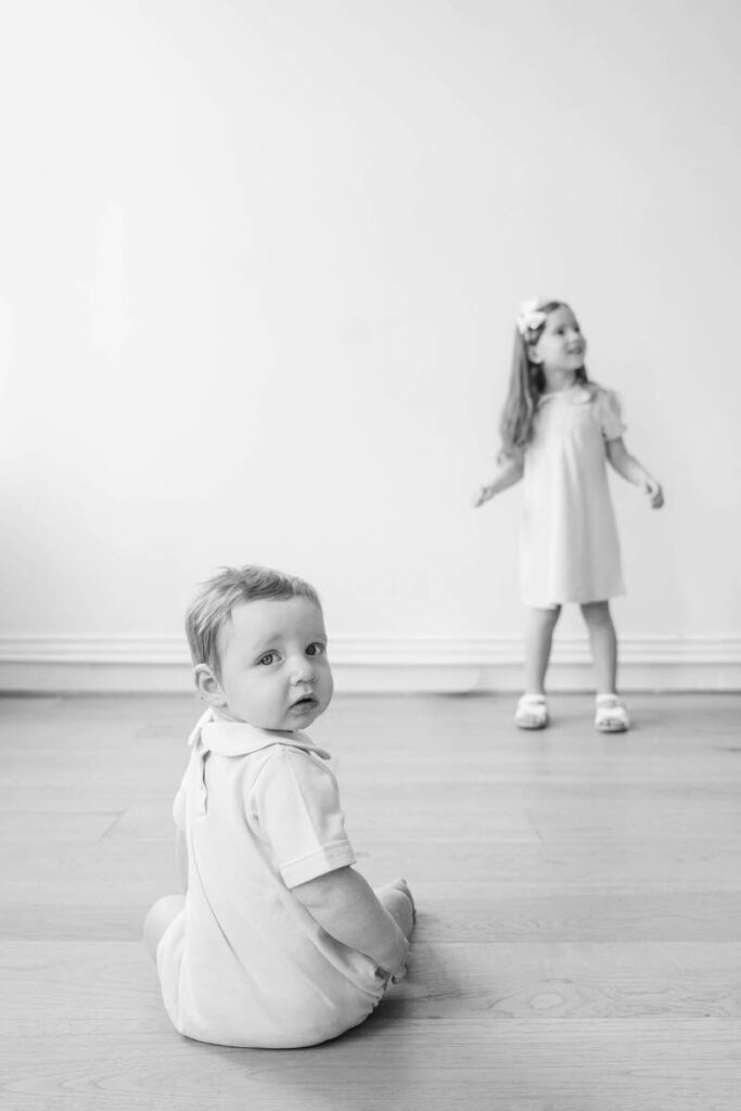 Baby sits and looks at the camera while his big sister plays in the background during his Louisville six month photos.