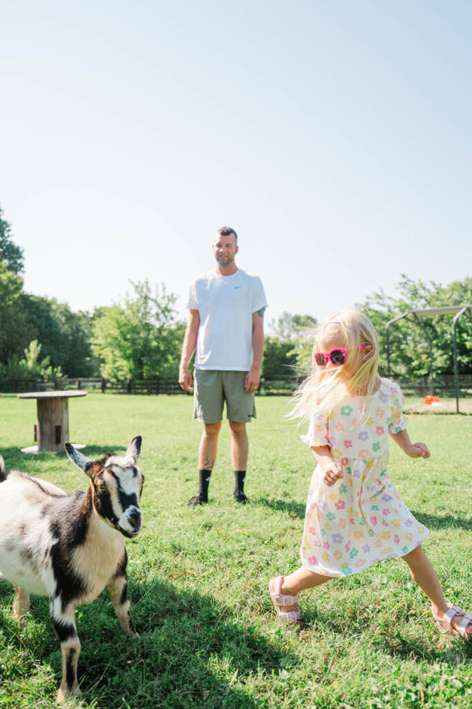 Little girl runs around her yard chasing a goat while her dad stands and laughs during their family session.