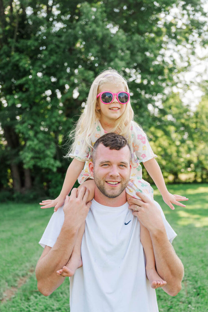 Little girl sits on dad's shoulders in their backyard, wearing sunglasses and smiling during their Louisville in-home family session.