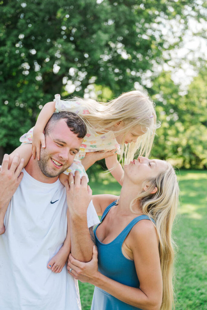 Little girl sits on dad's shoulders and reaches down to give her mom a kiss during their Louisville family session.