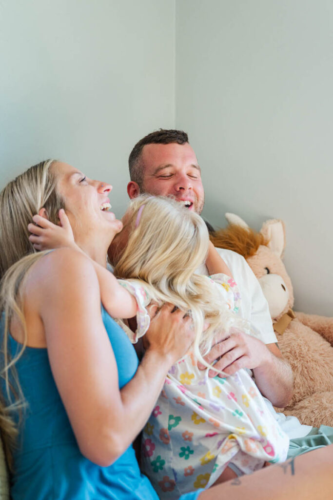 Little girl hugs her mom and dad on the bed while they laugh during their in-home family session.