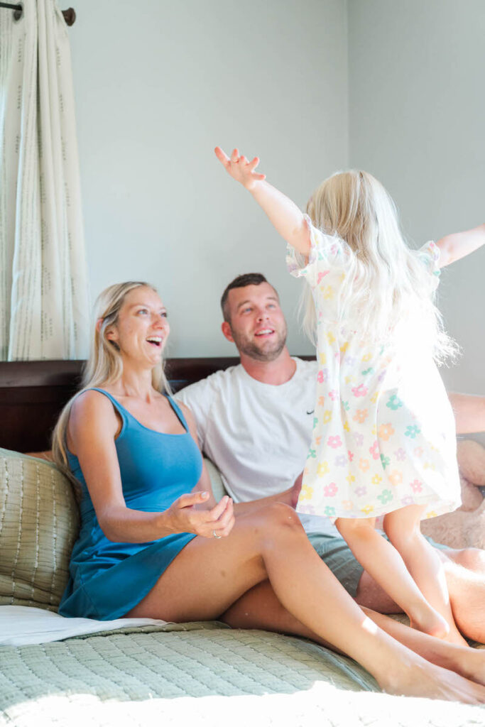 Little girl jumps on the bed into her mom and dad's arms during their in-home family session.