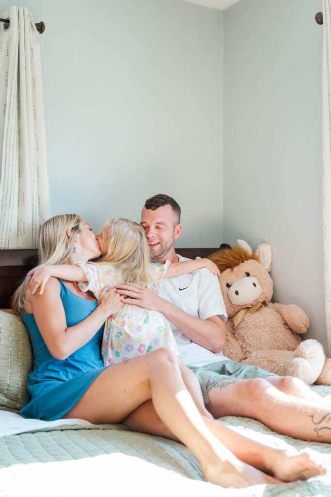 Little girl hugs her mom and dad while mom kisses her on the cheek during their in-home family session.