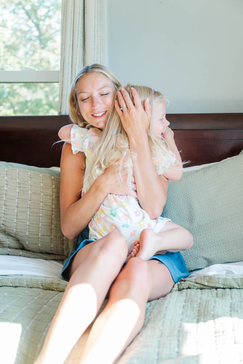 Little girl hugs mom on the bed while they both smile during their family photo session.