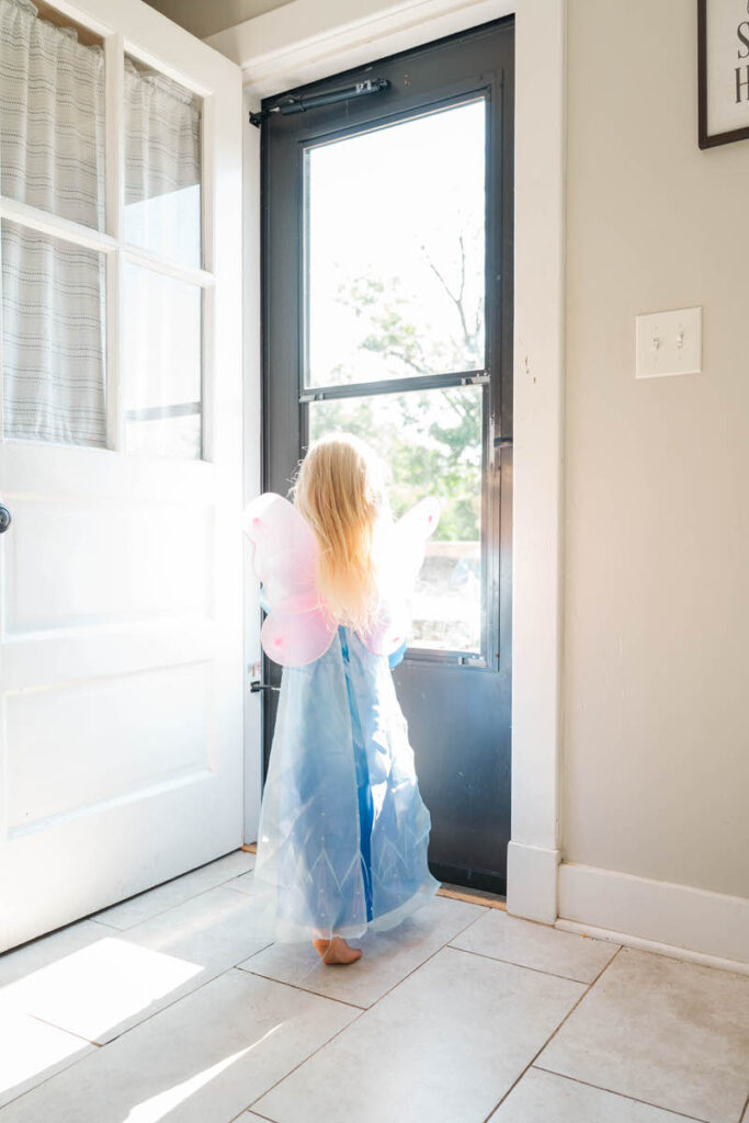 Little girl plays dress up, wearing butterfly wings in the sunshine of her home.