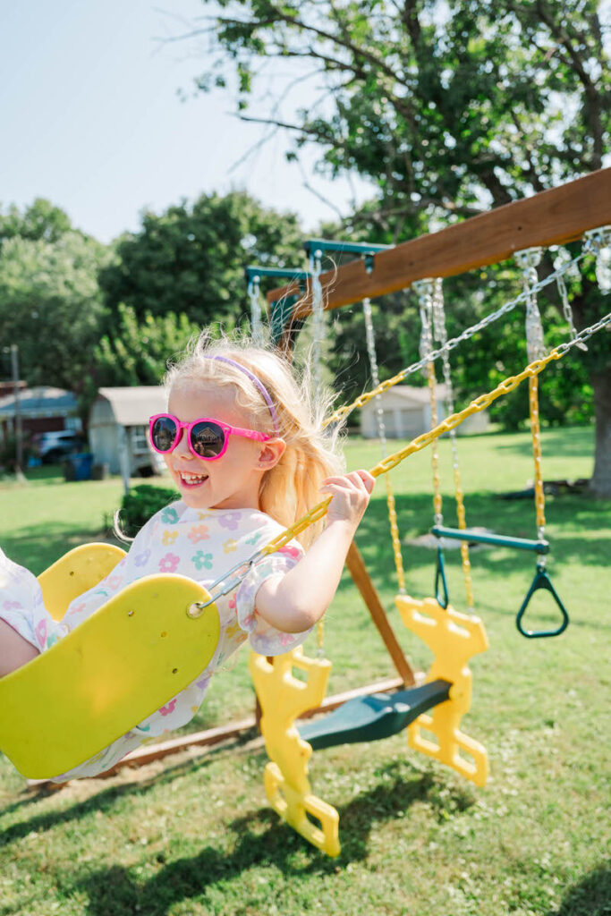 Little girl swings on the swing-set wearing sunglasses and smiling during her in-home family session.