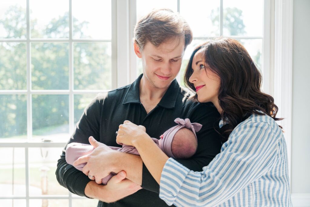 New parents smile while holding their newborn baby in front of a sunny window during their Louisville in-home newborn session.