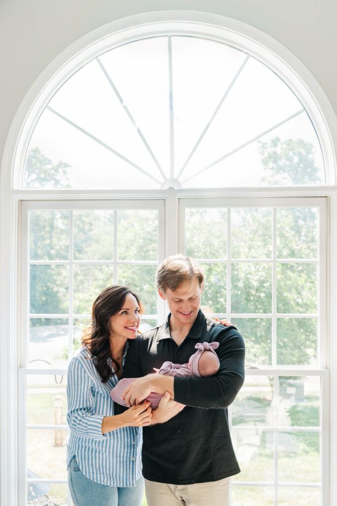 New parents smile while holding their newborn baby in front of a sunny window during their Louisville in-home newborn session.