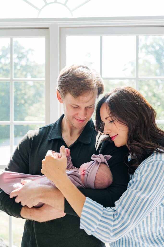 New mom and dad smile down at their baby girl in pretty sunlight during their Louisville in-home newborn session.
