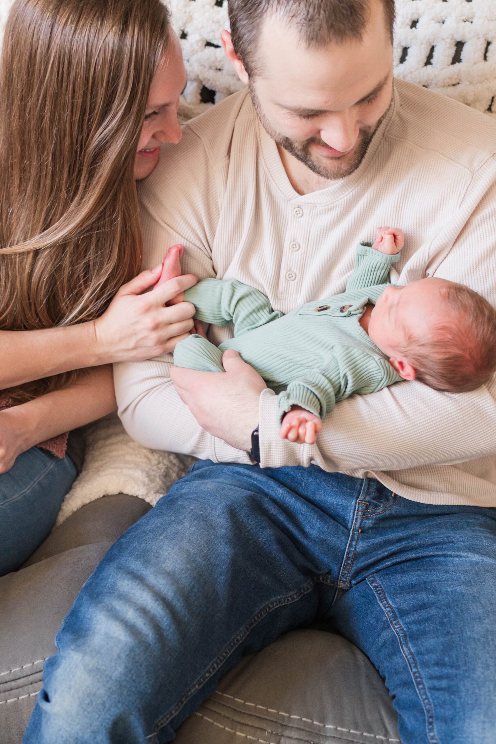 Mom and dad snuggle newborn baby on couch in their home.