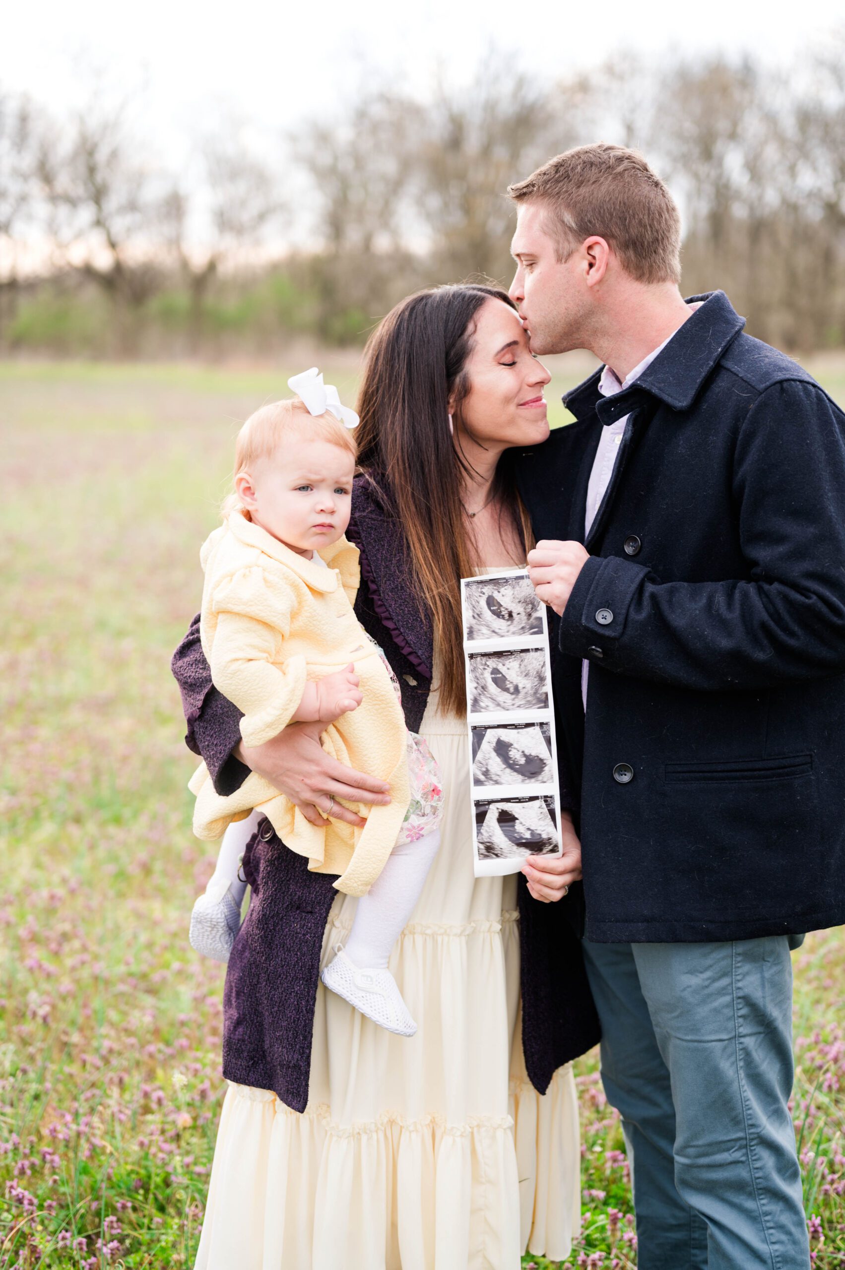 Louisville family stands together. Mom holding daughter, dad kissing her forehead. They hold an ultrasound for their pregnancy announcement session.