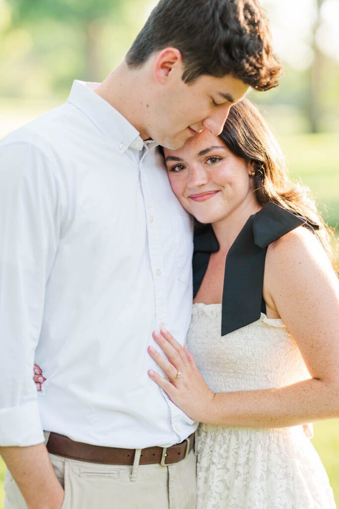 Bride-to-be smiles at the camera with her fiance looking down at her during their engagement session at Anchorage Trail