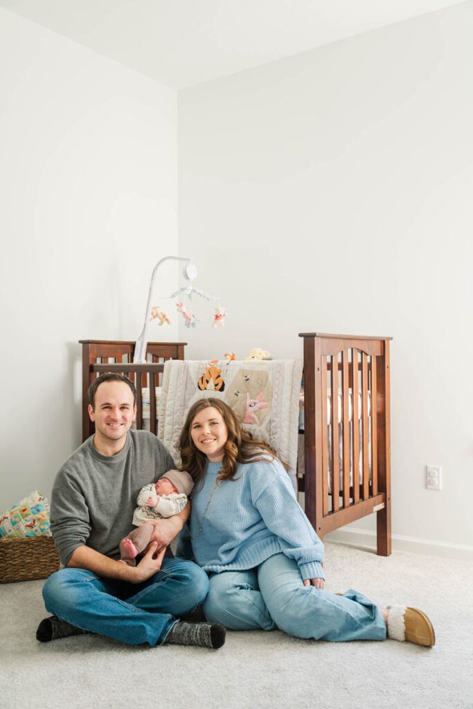 A mom, dad, and newborn sit on nursery floor.