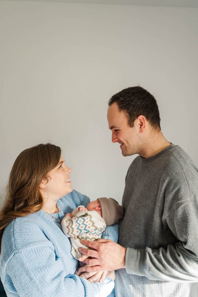 A new mom and dad smile at each other while holding their newborn baby.